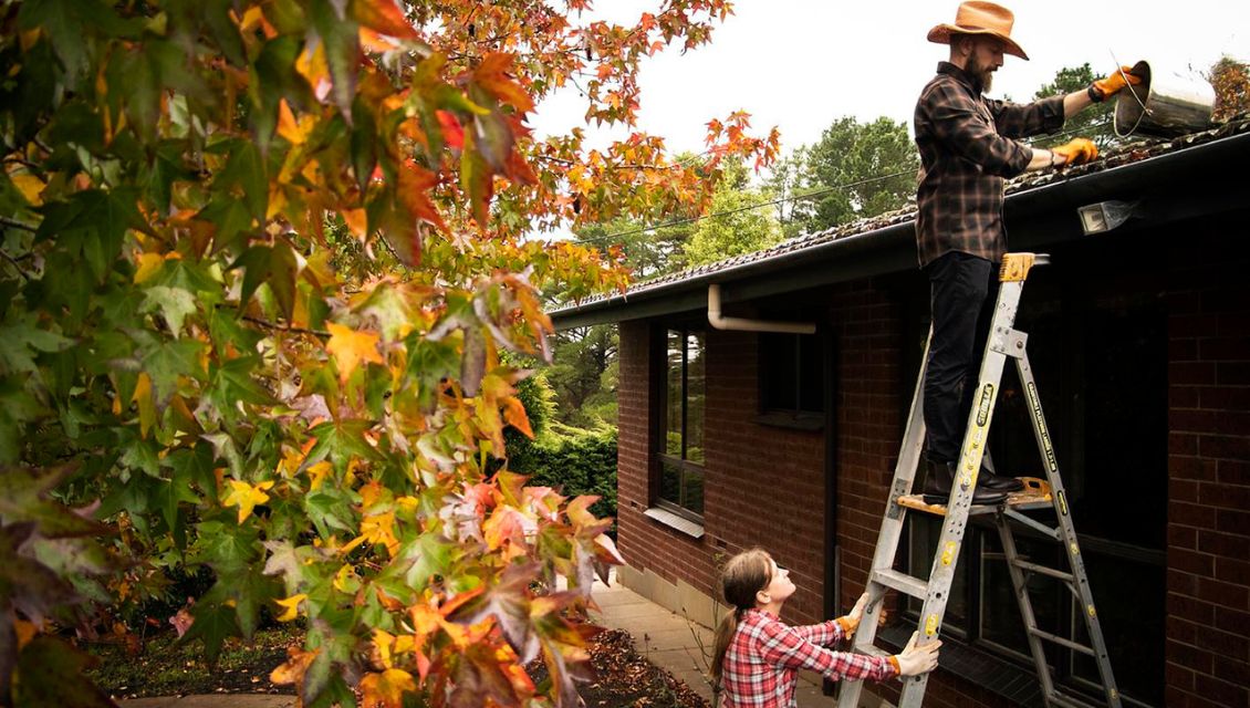 Image of a man on a ladder clearing gutters, a woman is holding the ladder stable.