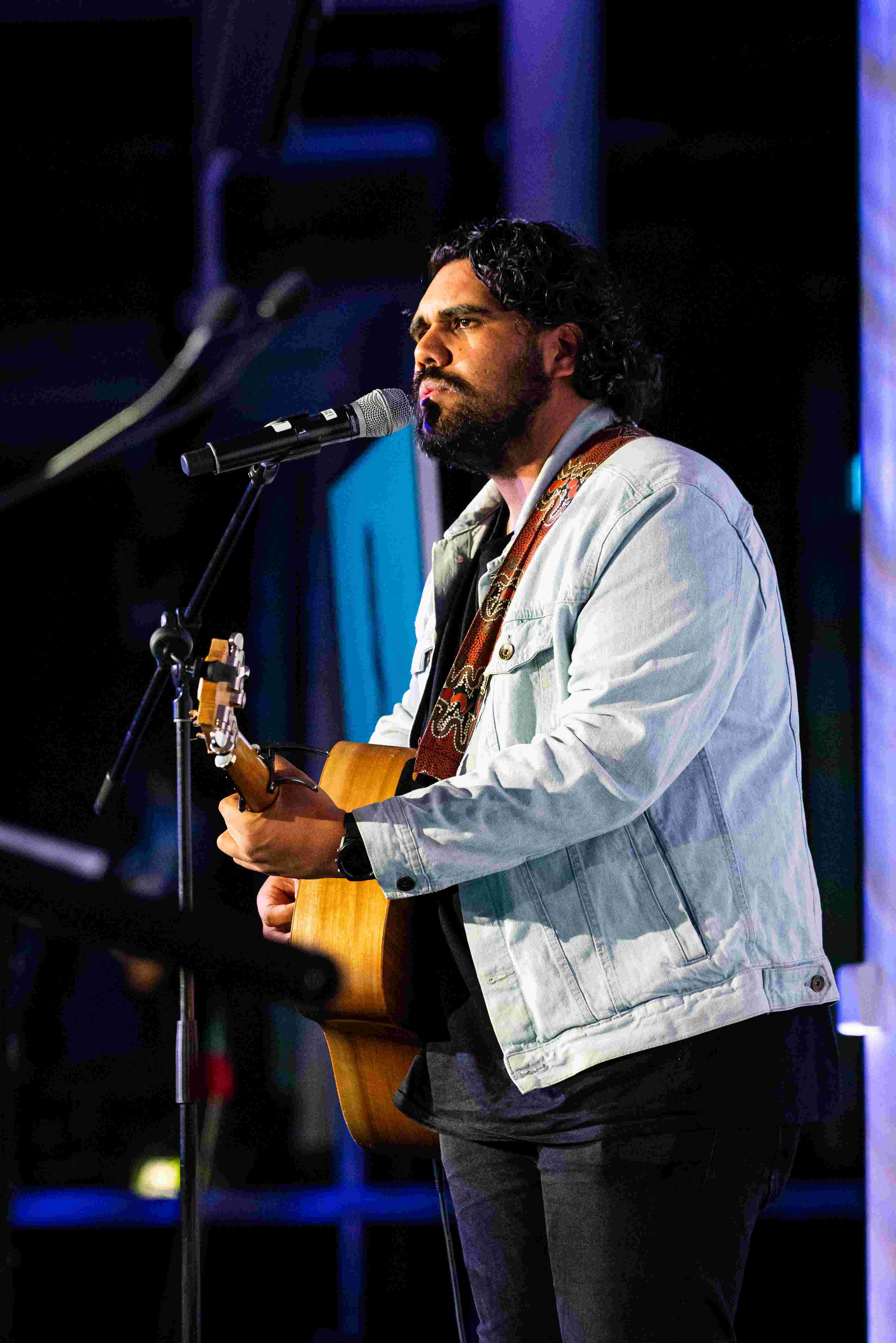 Image of Nathan May, he is performing on stage playing guitar and singing. He is wearing a denim jacket.