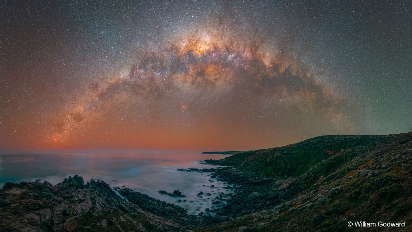 Image of the night sky, with stars appearing above a rocky coastline in an orange arc of galaxy.