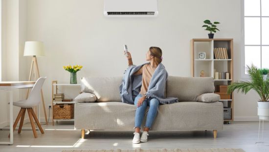 Image of a woman sitting on a couch with a blanket around her shoulders, holding the remote control to her airconditioner.