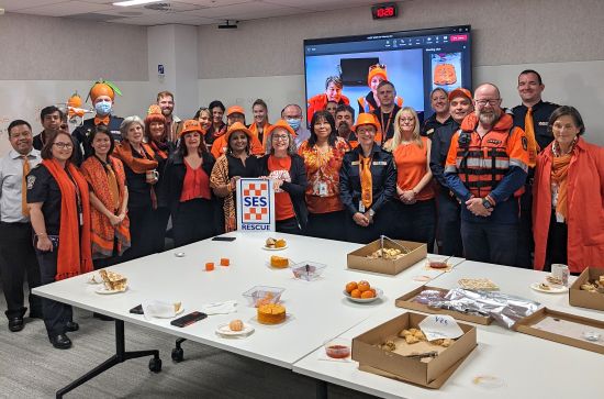 Image of a group of people wearing orange clothing and accessories, they are standing in front of a table with morning tea items.