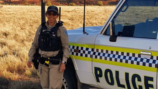 Image of Constable Alison Sampson wearing a police uniform and standing in front of a police vehicle. In the background are brown hills.