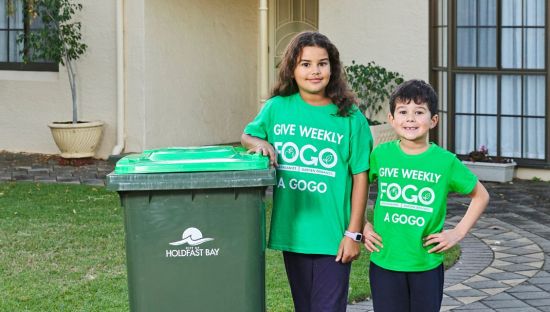 Image of two children, one boy and one girl, standing next a green-lidded bin. The children are wearing green t-shirts saying 'give weekly FOGO a gogo'.