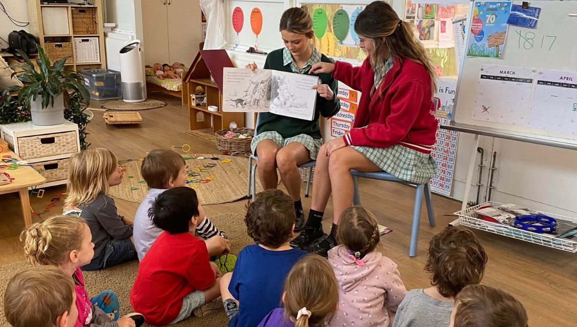Image of two high school aged girls sitting on chairs in front of a group of young daycare students, reading them a book.