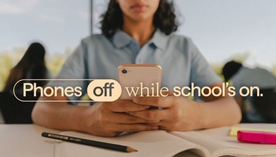 Image of female student holding mobile phone and sitting at a school desk. Text overlay reads: Phones off while school's on.