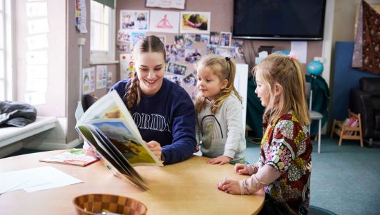Image of a teacher surrounded by preschool-aged children, they are in a circle and playing with musical instruments.