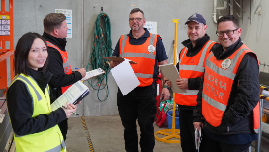 Image of five people wearing high vis vests at a work site and holding onto clipboards