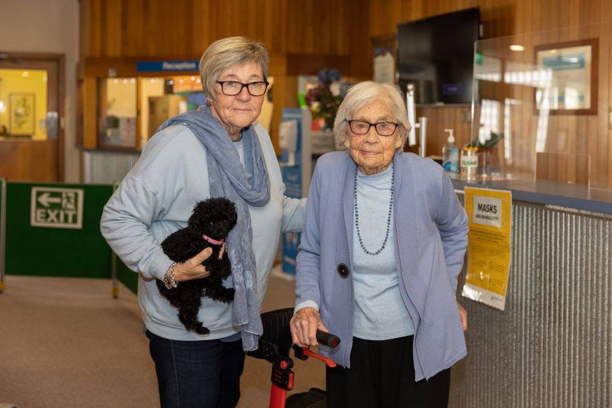 Image of two women, Jennie Jacobs is on the left holding a small black poodle puppy. Her mother is on the right, holding her walking frame.