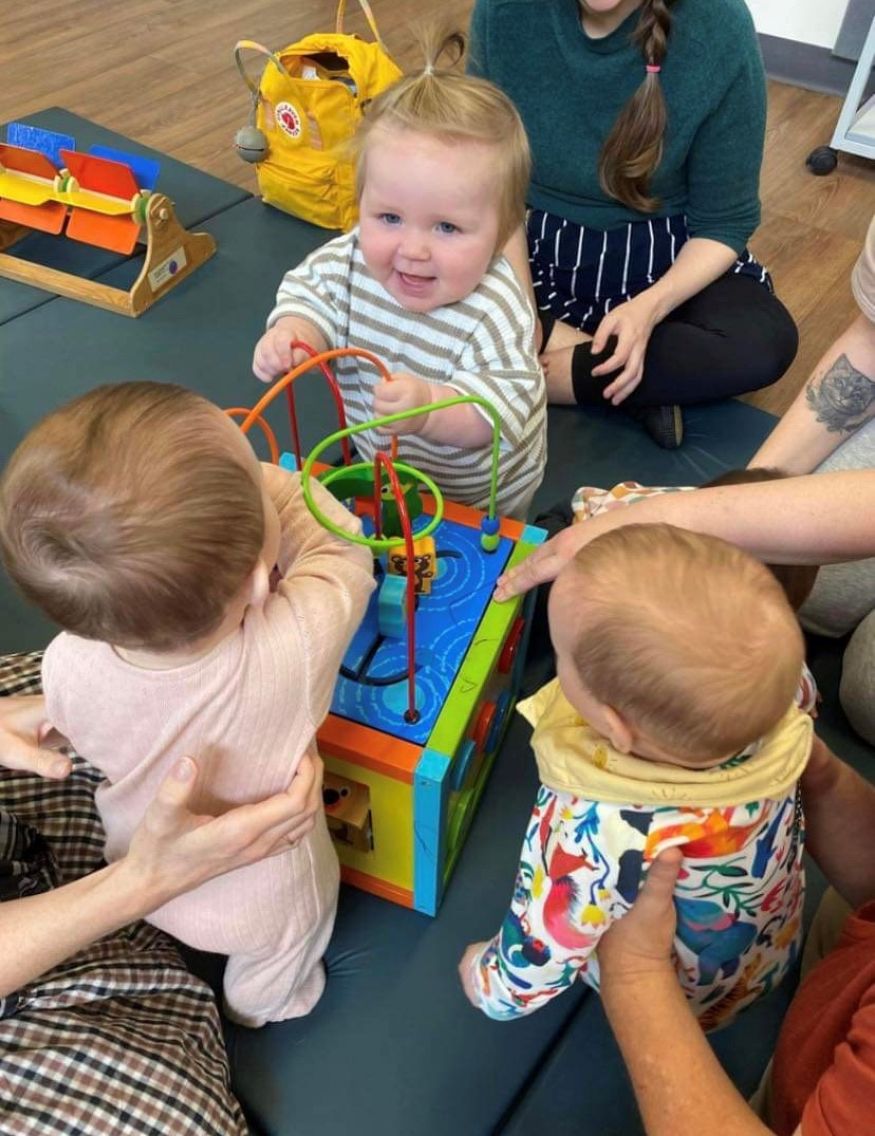 Three babies playing with toys and surrounded by their parents on a play mat