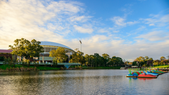 Image of the Torrens Lake with Adelaide Oval to the left, covered with trees and colourful paddle boats to the right.